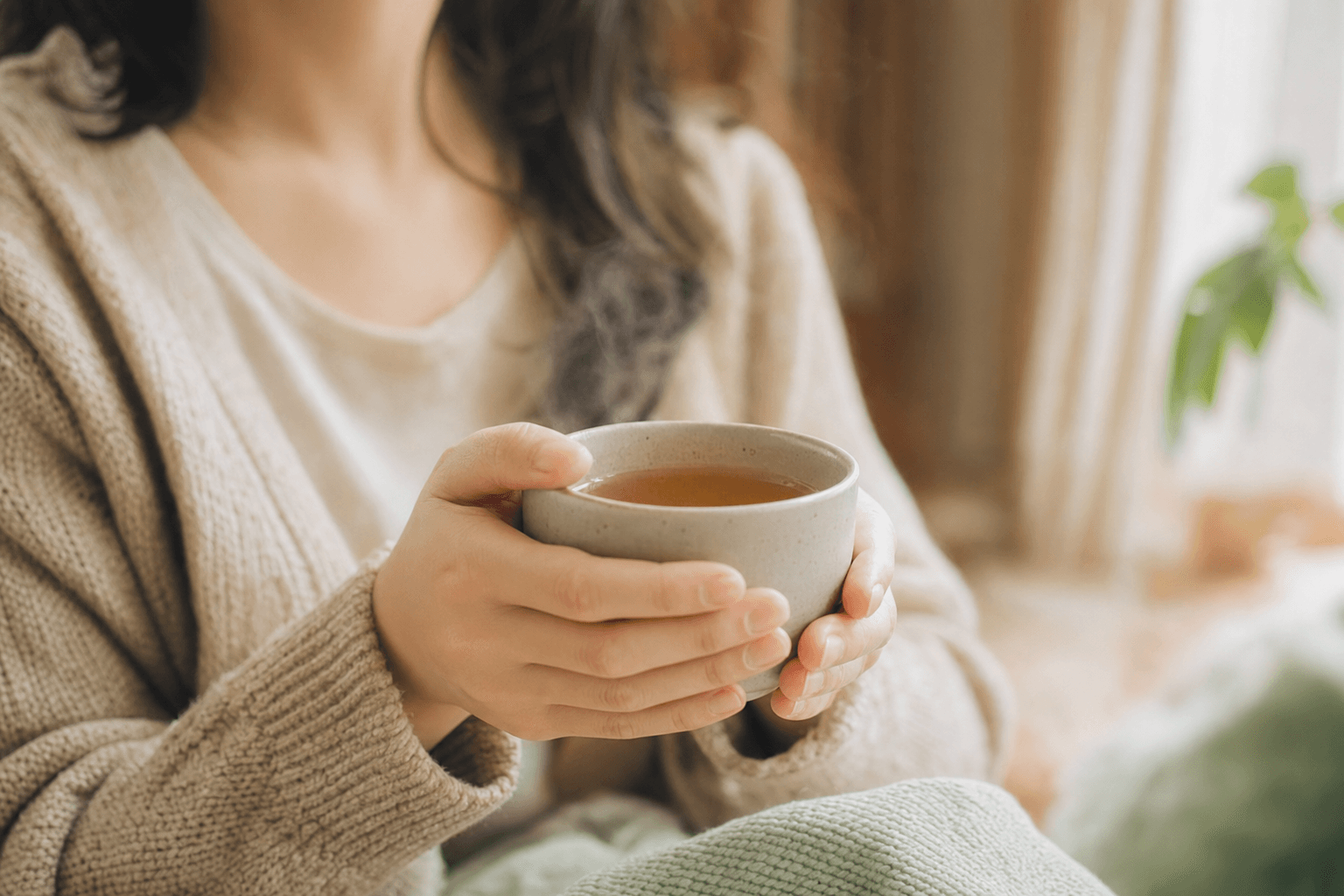 A woman relaxed at home cradling a simple warm cup of golden-brown Ember Crown Select Dark Oolong in both hands, at rest