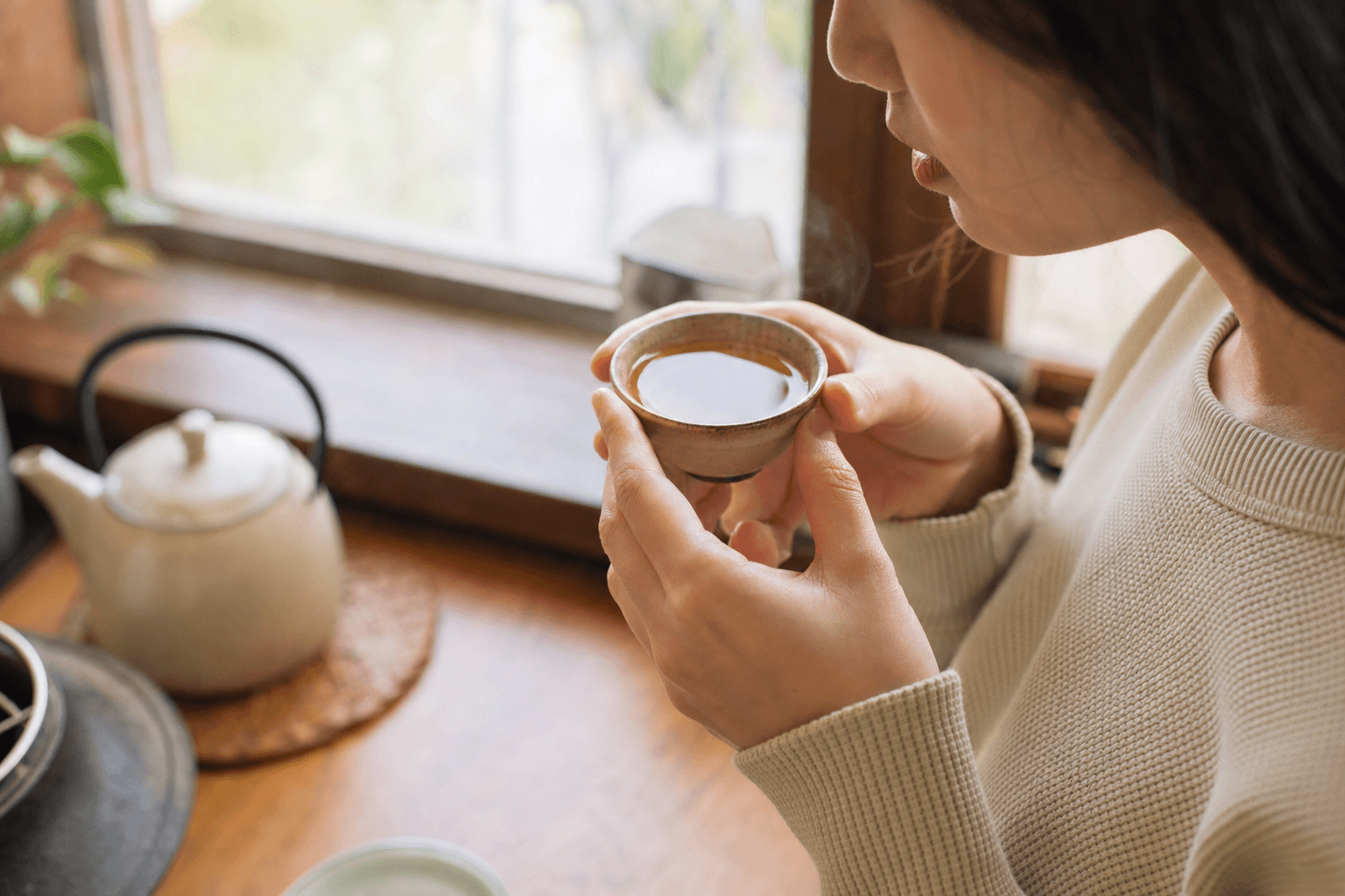 A young woman in a sunlit kitchen lifting a simple cup of golden-brown Ember Crown Select Dark Oolong, her face only partly visible