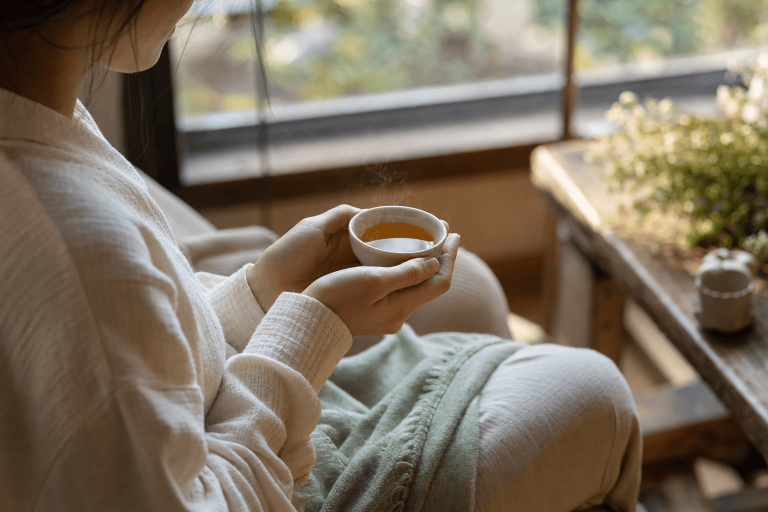 A young woman by a sunlit window cradling a small cup of golden-brown Ember Crown Signature Dark Oolong in both hands, at rest