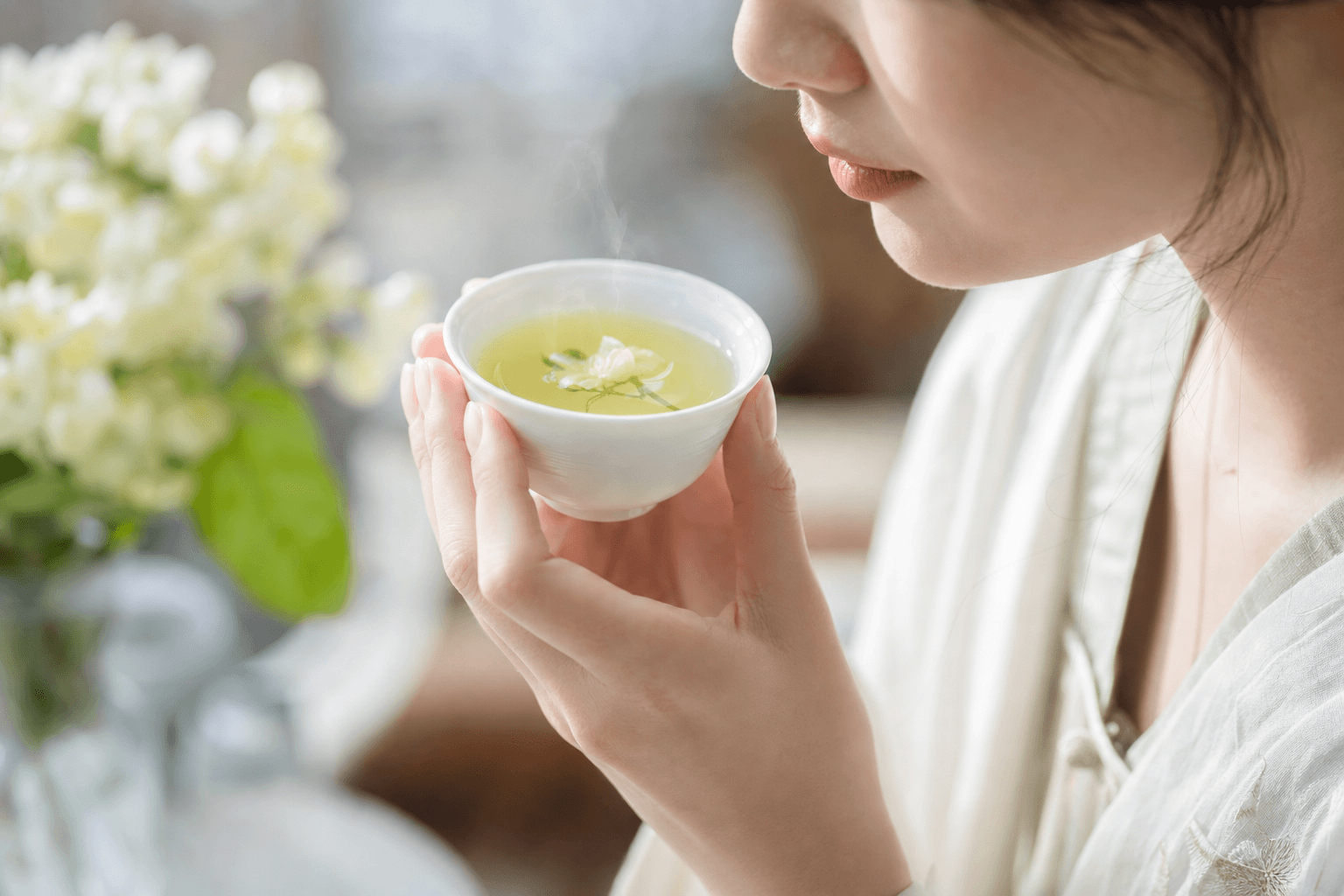 A woman bringing a small white porcelain cup of pale yellow-green Huatan Reserve Jasmine up to savour it, her face only partly visible, jasmine blossoms nearby