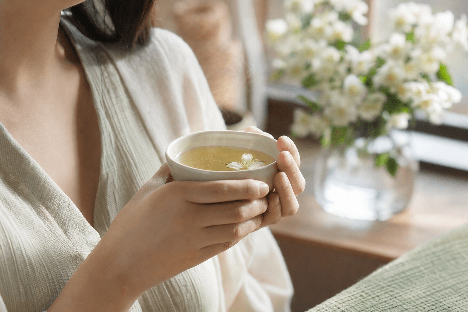A woman relaxed at home cradling a simple cup of pale Huatan Select Jasmine in both hands, at rest, with jasmine blossoms nearby