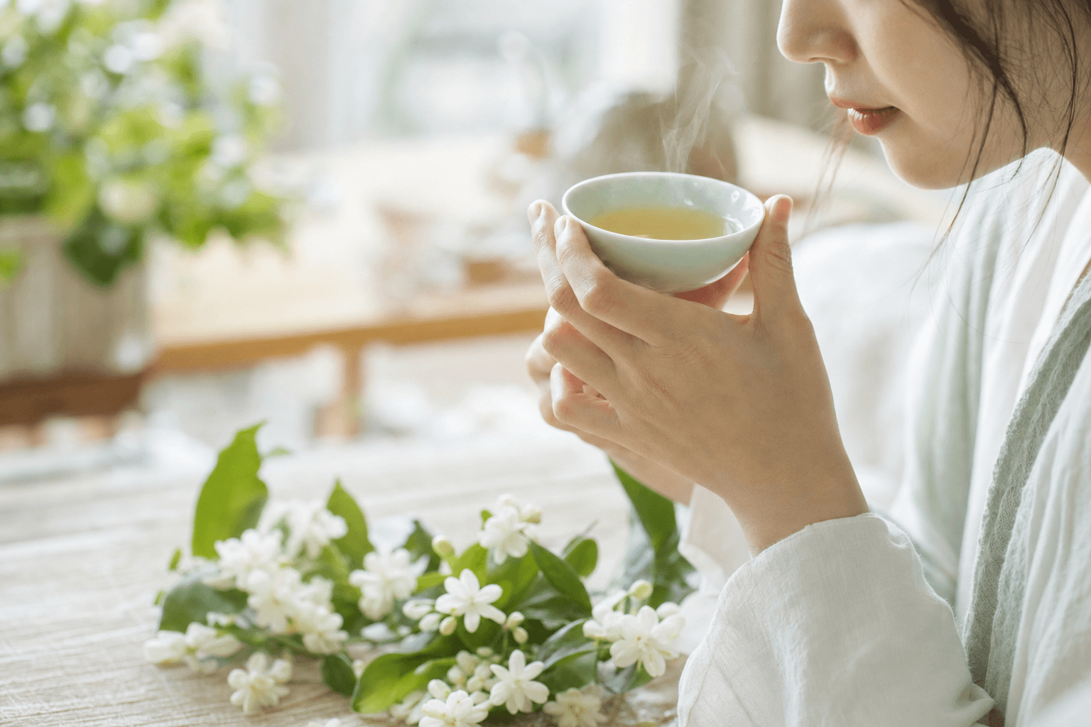A young woman in a cozy room sipping a small cup of pale yellow-green Huatan Signature Jasmine, the cup cradled in both hands, her face only partly visible