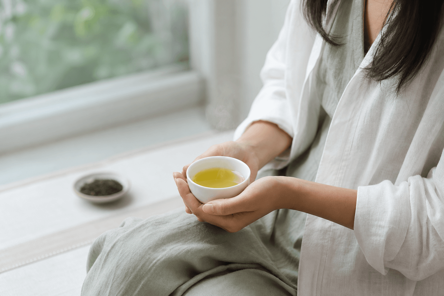 A woman by a bright window cradling a small cup of yellow-green Tie Guan Yin oolong, at rest