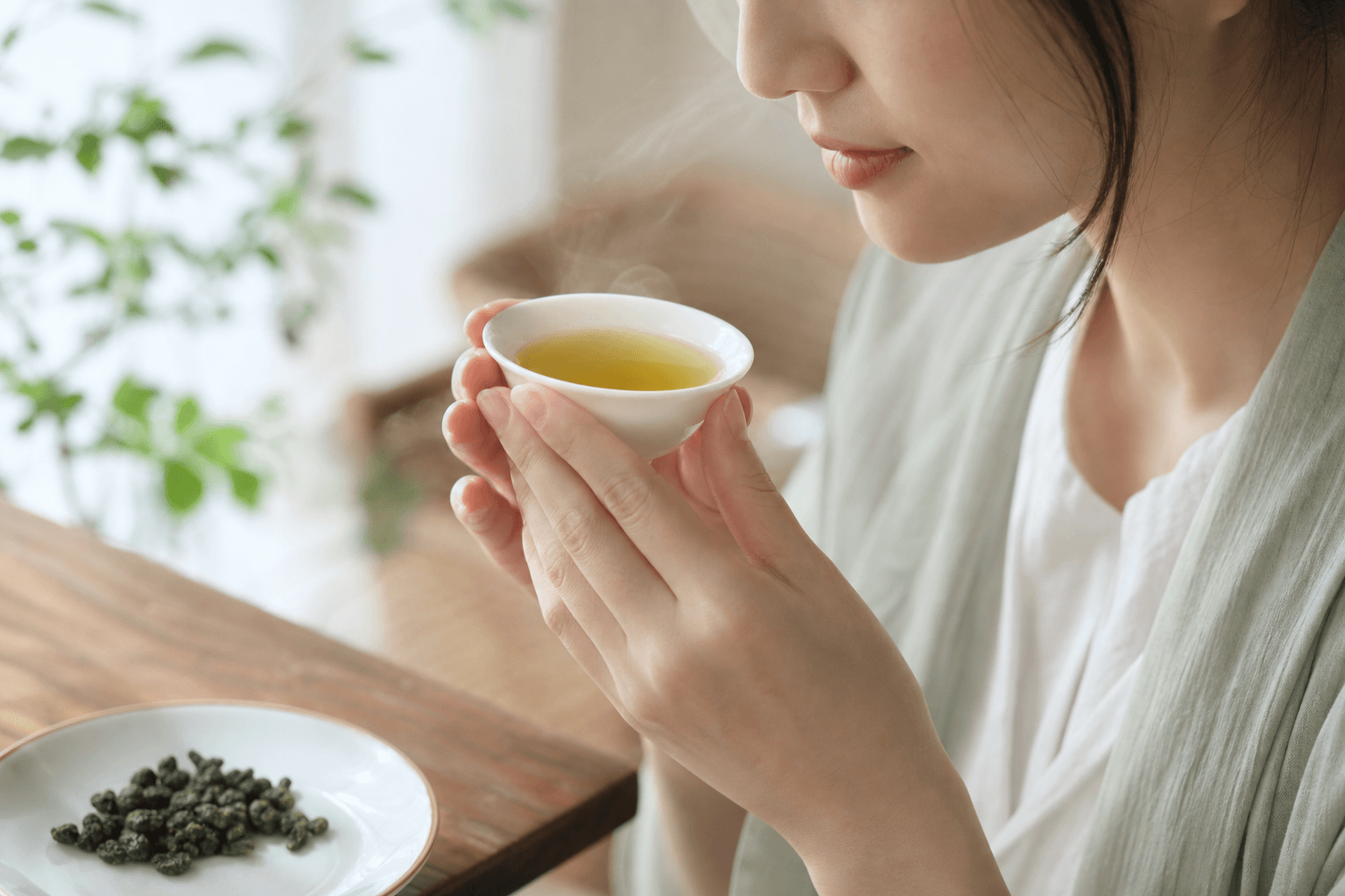 A woman bringing a small white porcelain cup of clear yellow-green Tie Guan Yin oolong up to savour the aroma, her face only partly visible