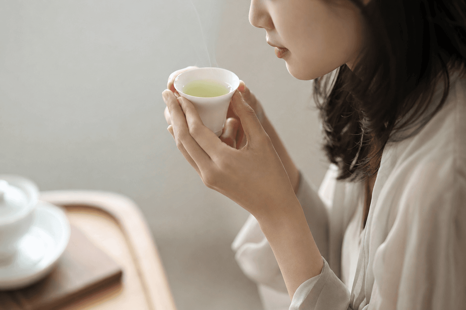 A poised woman bringing a small white porcelain cup of pale jade Mountain Crown Reserve Oolong up to savour it, her face only partly visible