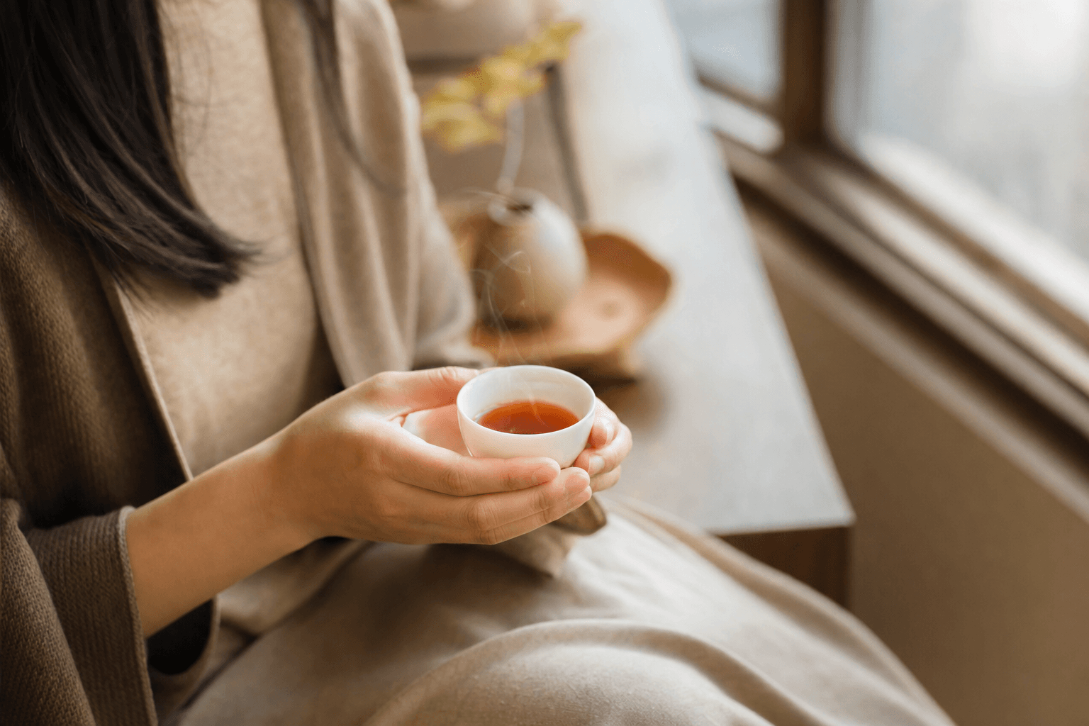 A poised woman by a bright window cradling a small white porcelain cup of clear reddish-brown Yunnan Crown Reserve Black Tea, at rest