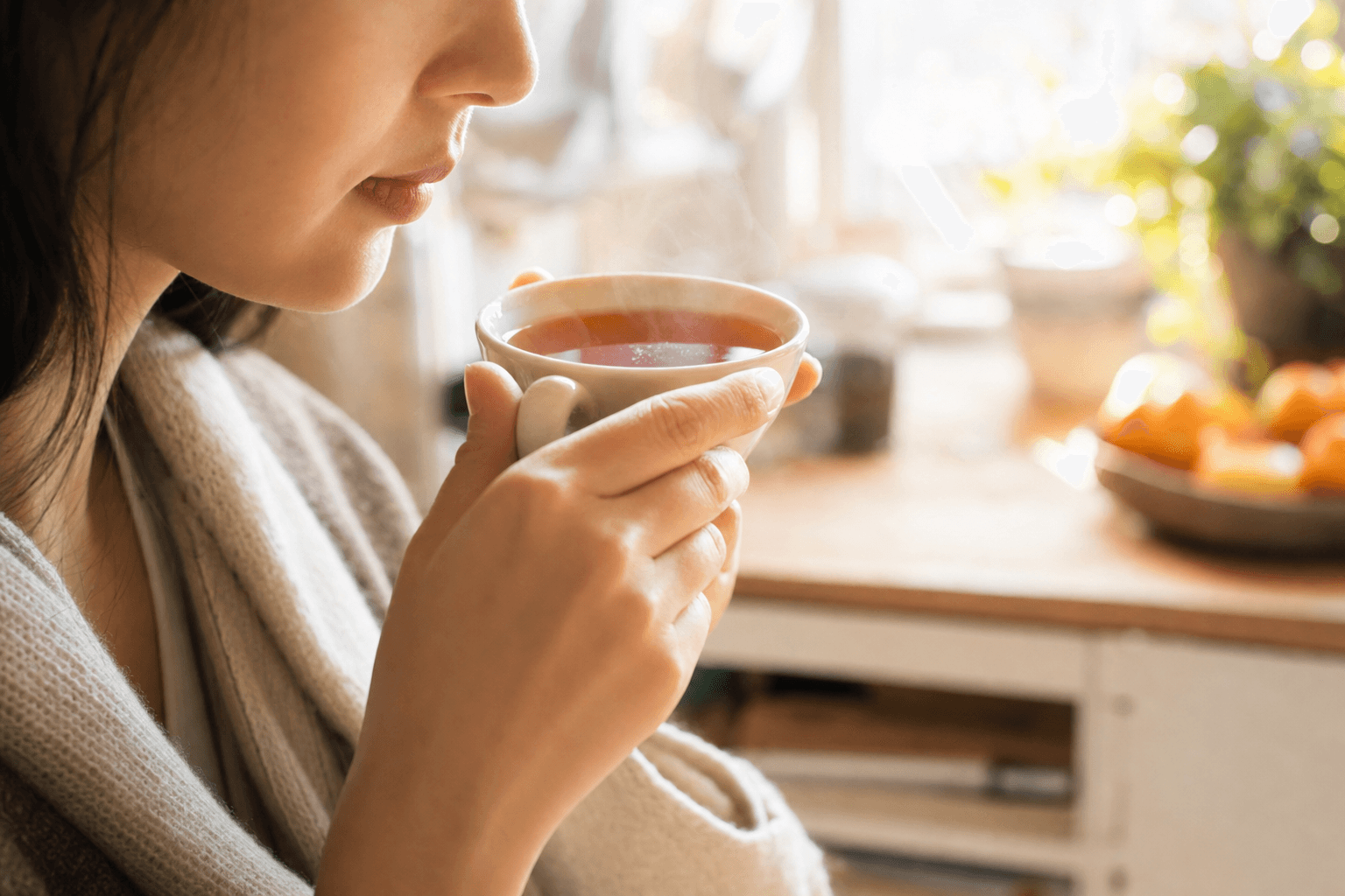 A young woman in a sunlit kitchen lifting a simple cup of clear reddish-brown Yunnan Crown Select Black Tea, her face only partly visible