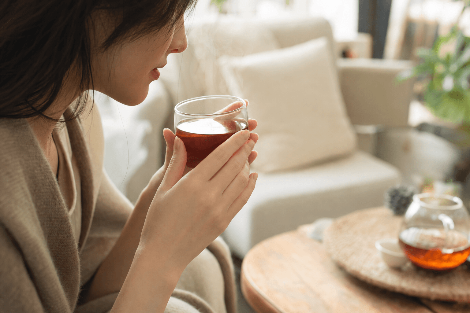A young woman in a cozy room sipping a small cup of clear reddish-brown Yunnan Crown Signature Black Tea, the cup cradled in both hands, her face only partly visible