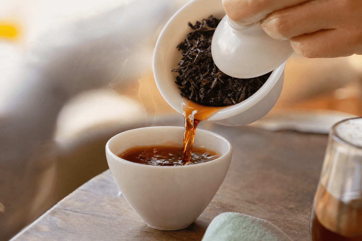Dark reddish-brown Yunnan Crown Signature Pu-erh poured from a gaiwan into a small cup on a warm wooden table, steam rising