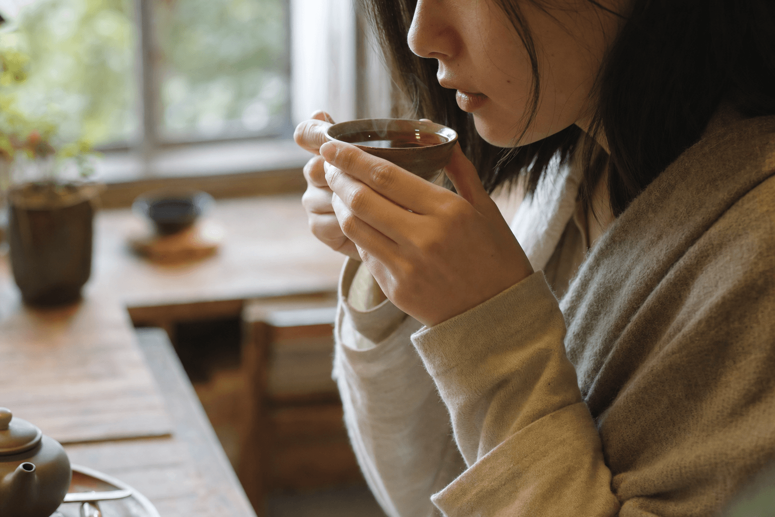 A young woman in a cozy room sipping a small cup of dark reddish-brown Yunnan Crown Signature Pu-erh, the cup cradled in both hands, her face only partly visible