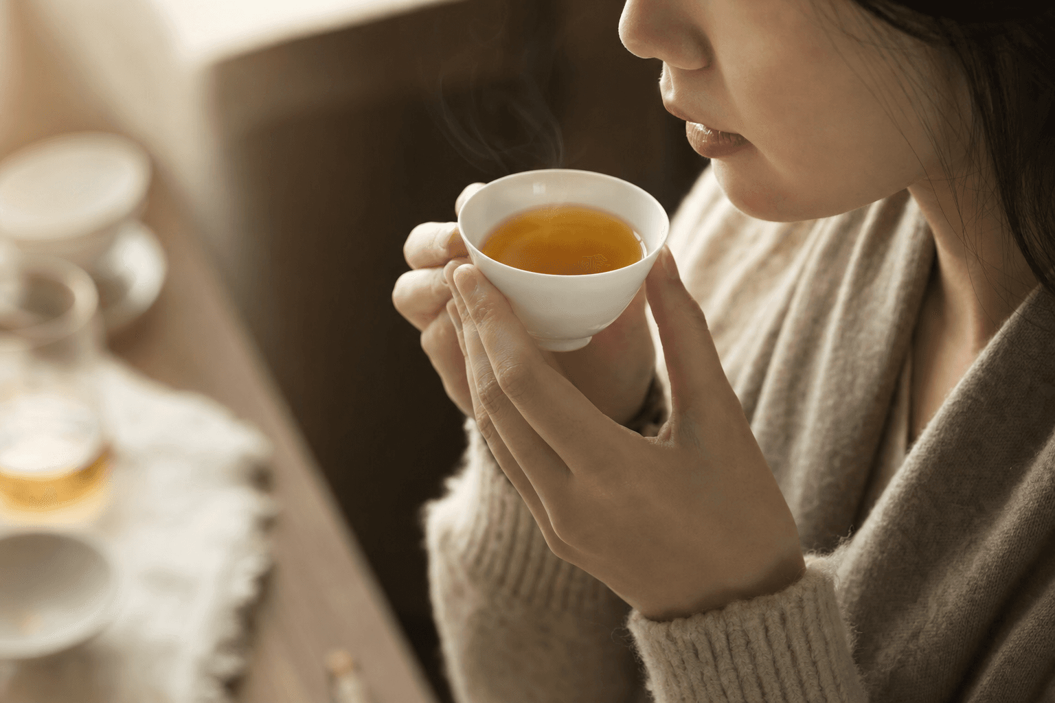 A poised woman bringing a small white porcelain cup of golden-brown Ember Crown Reserve Dark Oolong up to savour it, her face only partly visible