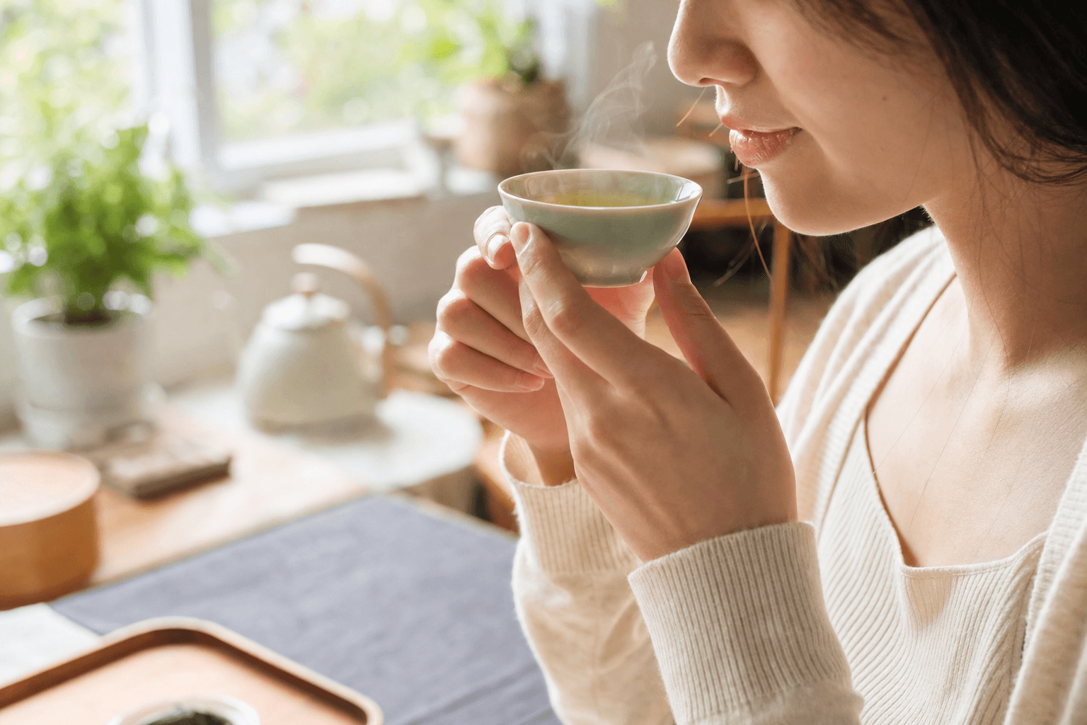A young woman in a sunlit morning kitchen lifting a small cup of pale jade Mountain Crown Select Oolong for the first sip of the day, her face only partly visible