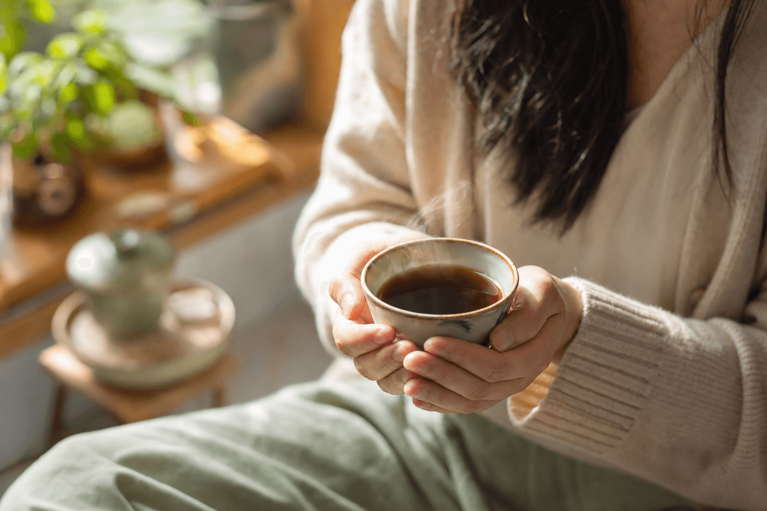 A woman relaxed at home cradling a simple warm cup of dark reddish-brown Yunnan Crown Select Pu-erh in both hands, at rest